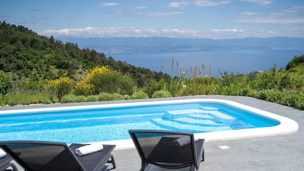 A swimming pool with lounge chairs, surrounded by greenery and a view of the sea.