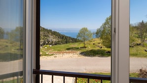 A view from a window overlooking a road with a grassy area and trees, leading to a body of water and distant hills.