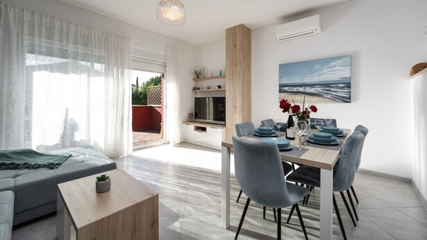Ein modernes Wohnzimmer mit einer Couch, einem Esstisch und Stühlen. Es gibt einen Fernseher, einen kleinen Tisch mit einer Pflanze und ein Bild an der Wand.