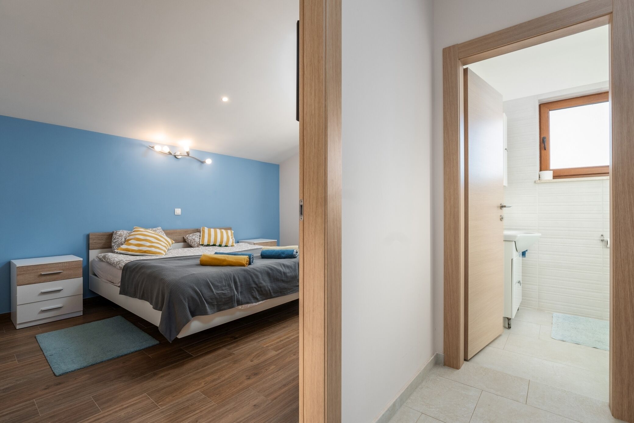 A bedroom with a bed, bedside table, and a nightstand. There is a bathroom visible through an open door.