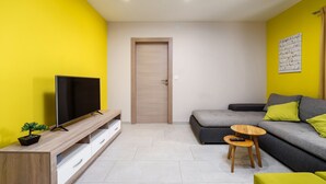 A modern living room with a flat-screen TV on a wooden TV stand, a grey sofa with green cushions, a small wooden table, and a door in the center. - Apartment Ulika (Istria - Visnjan) (Visnjan)