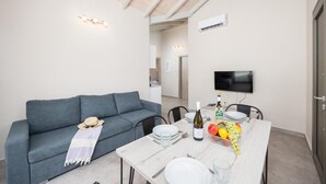 A modern living room with a grey sofa, a dining table set with plates, glasses, and a bottle of wine. A television is mounted on the wall, and there is a ceiling fan.