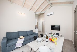 Een moderne woonkamer met een grijze bank, een eettafel gedekt met borden, glazen en een fles wijn. Een televisie is aan de muur bevestigd en er is een plafondventilator.