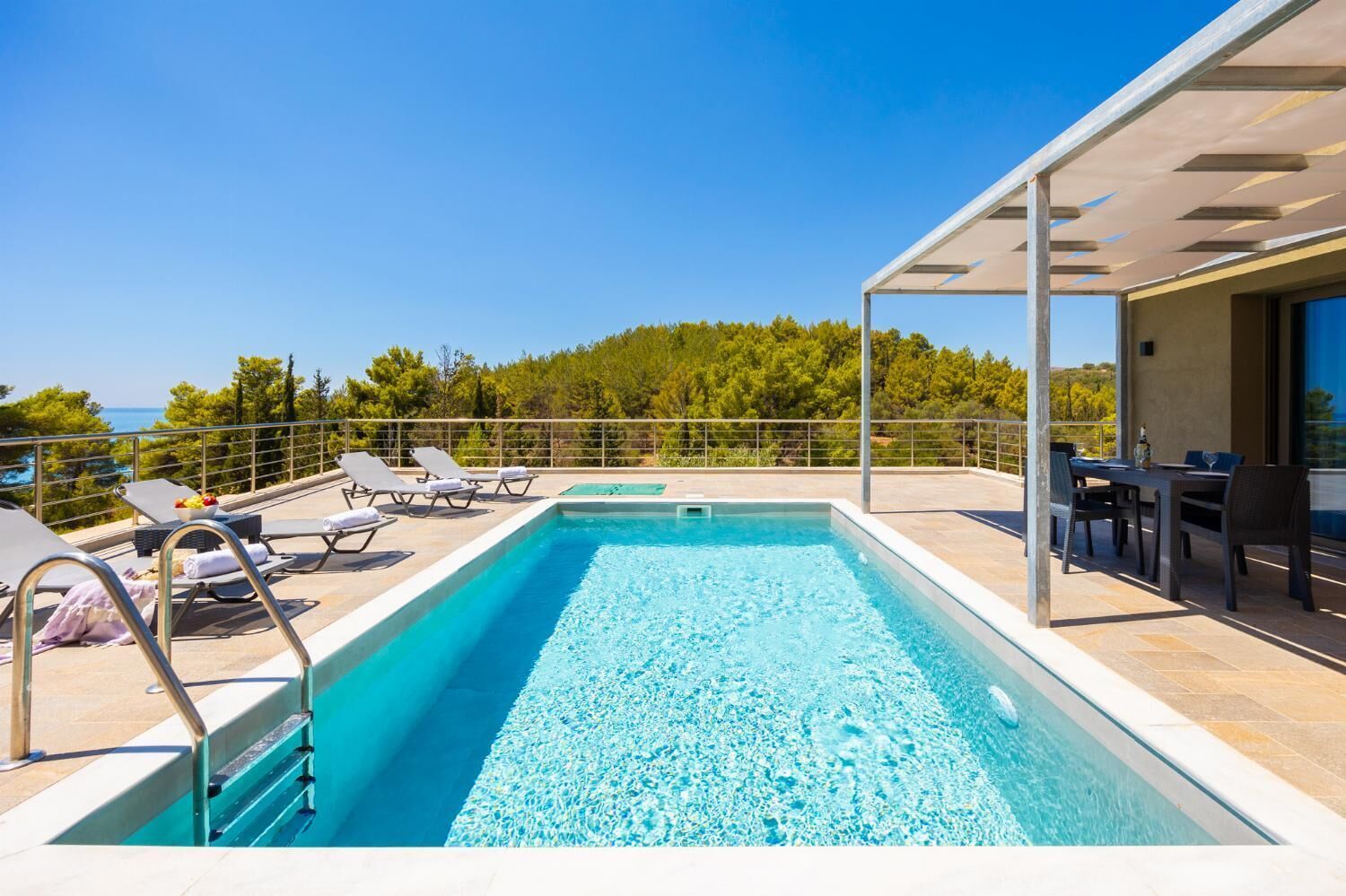 Une piscine sur le toit, dotée de chaises longues et d’un espace repas, le tout entouré d’arbres et surplombant un ciel dégagé.