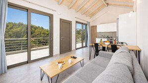 Ein modernes Wohnzimmer mit einer Couch, einem Esstisch mit Stühlen, einer Küchenzeile und großen Schiebeglasüren zum Balkon mit Blick auf Bäume.