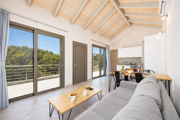 Een moderne woonkamer met een bank, een eettafel met stoelen, een keukenhoek en grote schuifdeuren die uitkomen op een balkon met uitzicht op bomen.