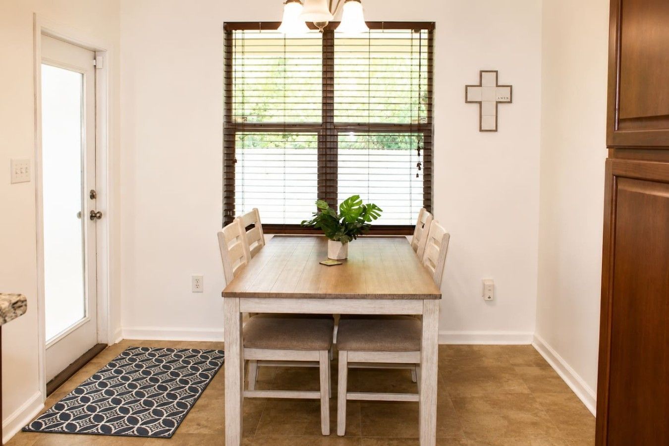 A dining area with a wooden table, four chairs, a potted plant, and a window with blinds.