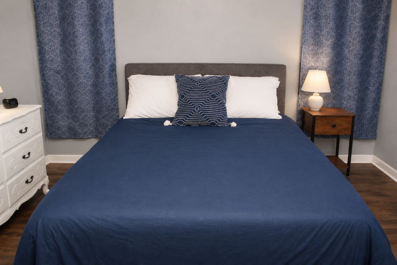 A bedroom with a large bed, two pillows, a bedside table with a lamp, and a dresser.