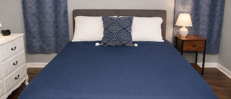 Quarto com uma cama grande, duas almofadas, mesa de cabeceira com luminária e cômoda.