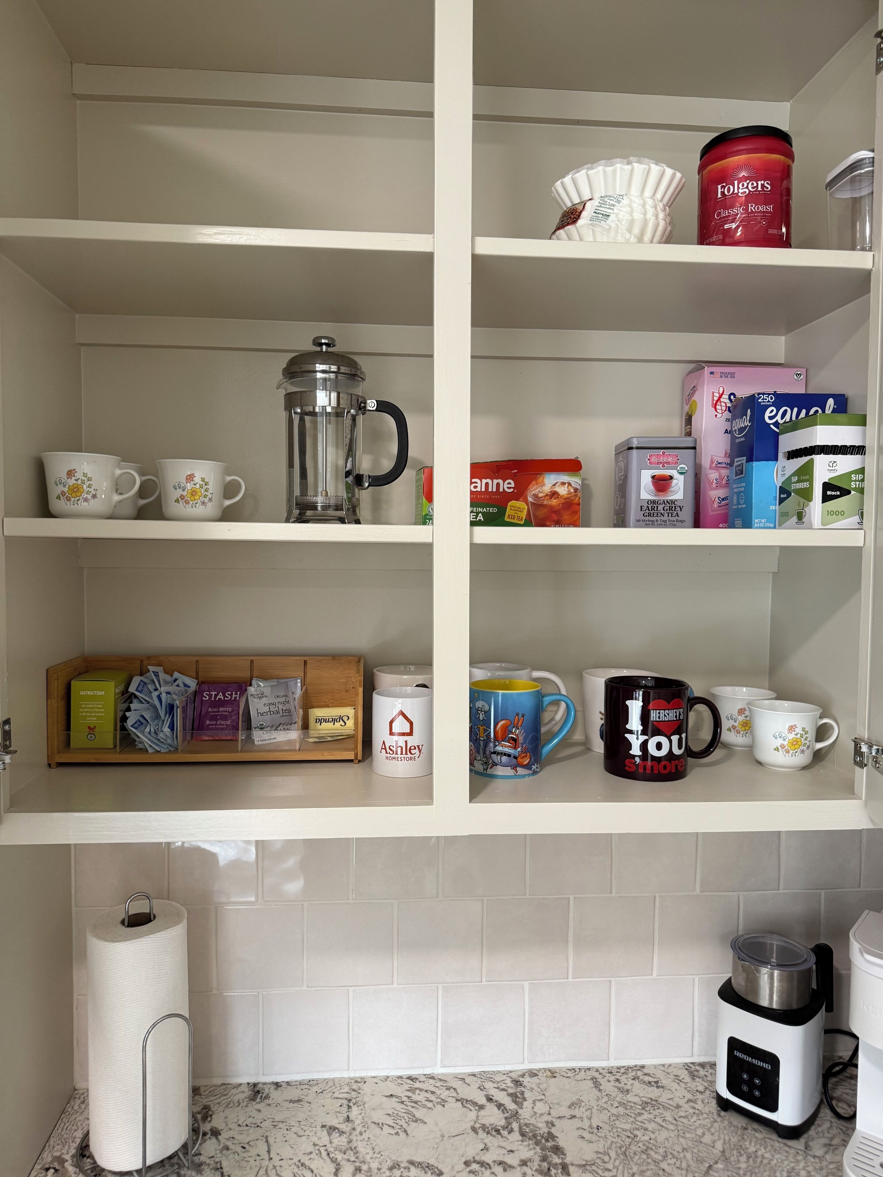 A kitchen with white shelves containing various items like a coffee maker, tea boxes, and mugs.