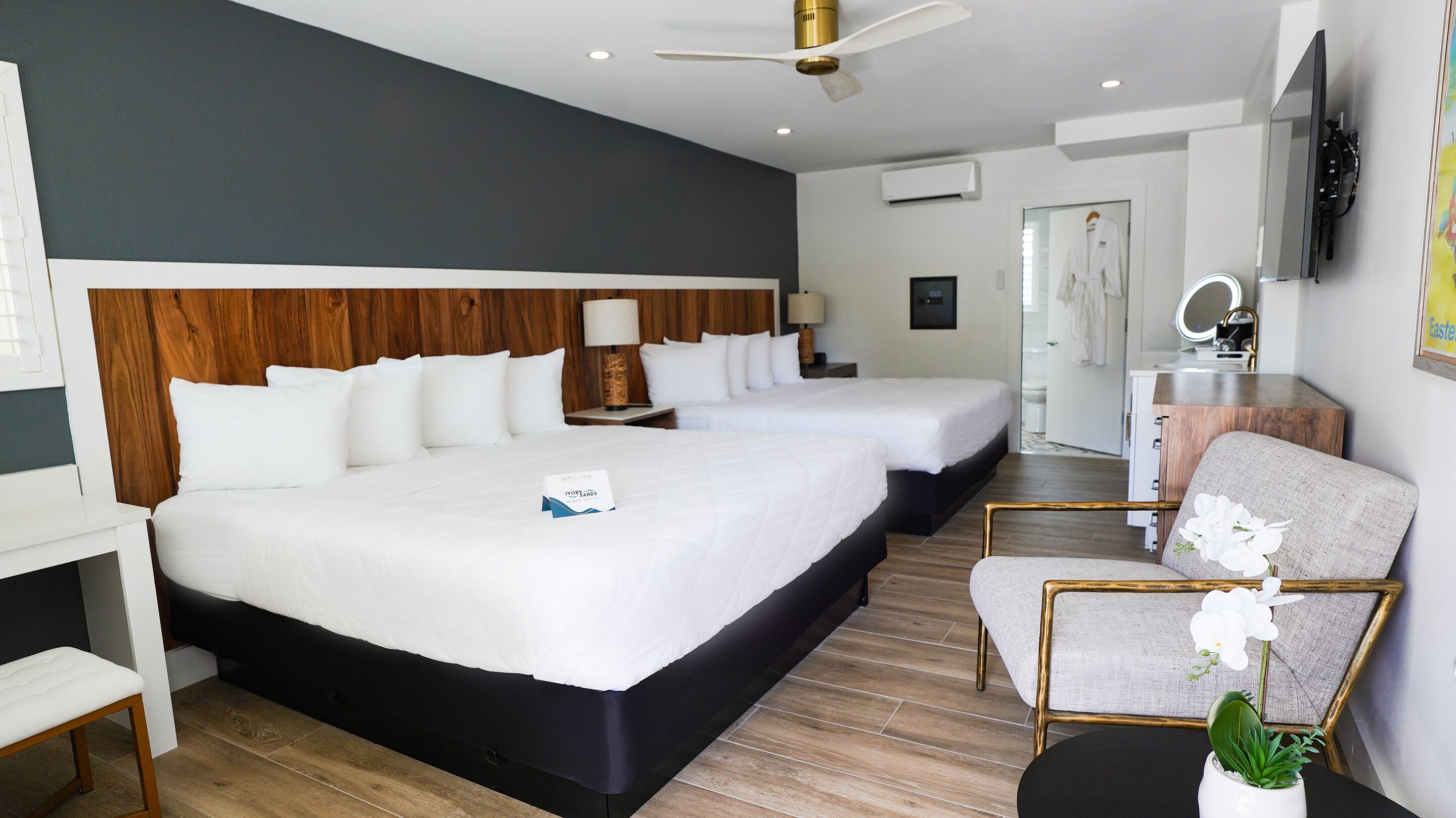 A hotel room with two beds, a desk, a chair, a small table with a plant, and a bathroom visible through an open door.