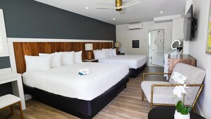 Habitación de hotel con dos camas, un escritorio, una silla, una mesita con una planta, y un baño visible a través de una puerta abierta.