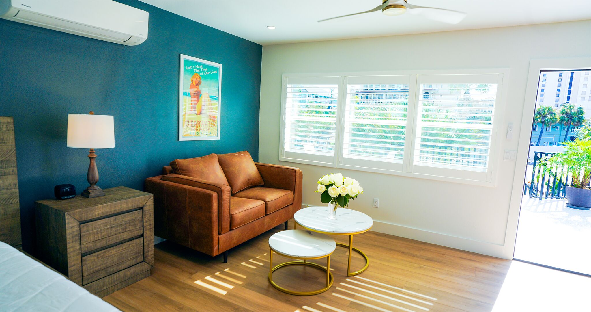 A living room with a brown leather sofa, a small round table with a vase of white flowers, a wooden side table with a lamp, and a large window with blinds.