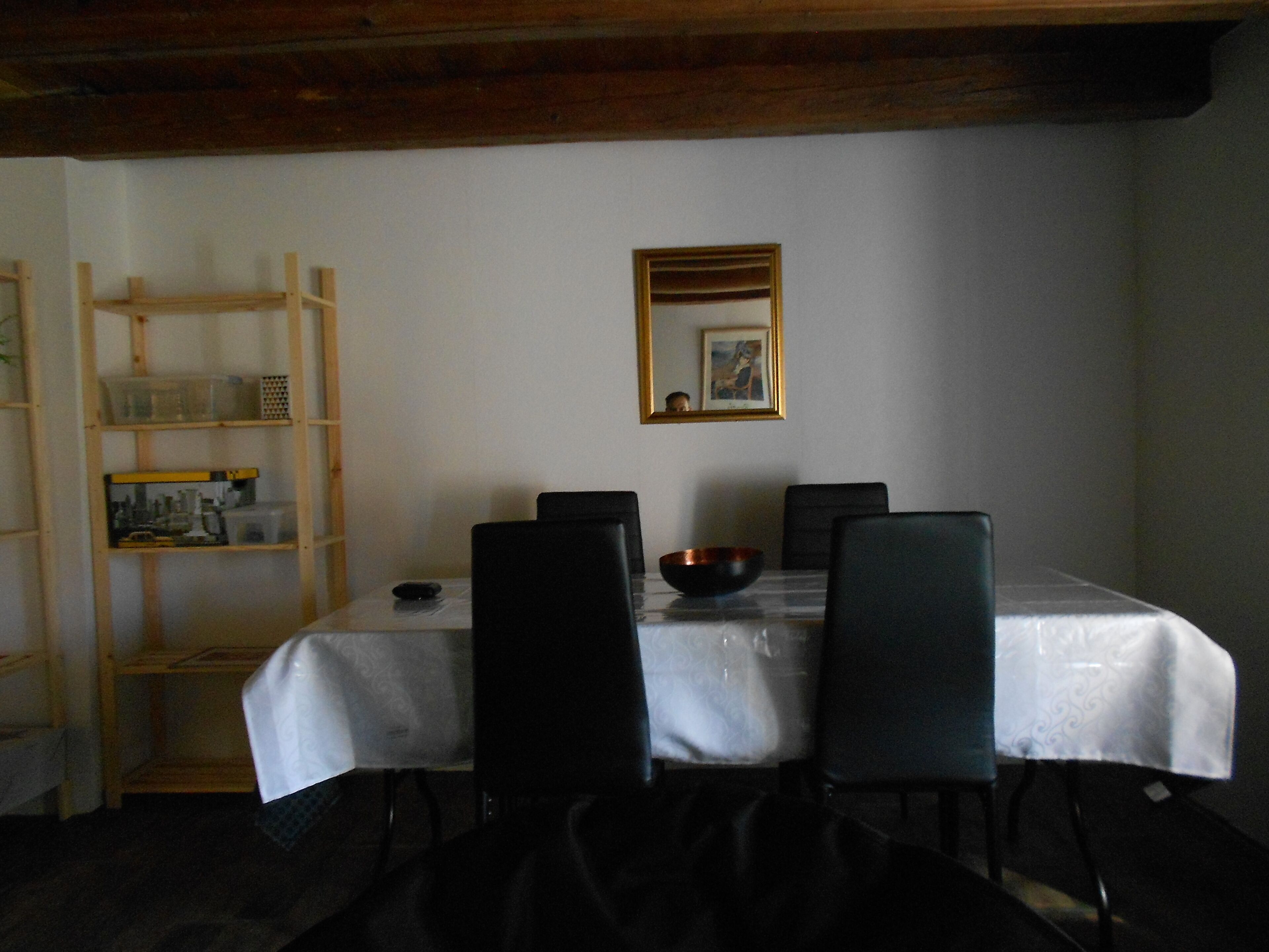 A dining area with a table covered by a white tablecloth, surrounded by four black chairs. There is a wooden shelf unit to the left with various items on it. A framed picture hangs on the wall above the table.