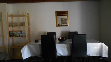 A dining area with a table covered by a white tablecloth, surrounded by four black chairs. There is a wooden shelf unit to the left with various items on it. A framed picture hangs on the wall above the table.