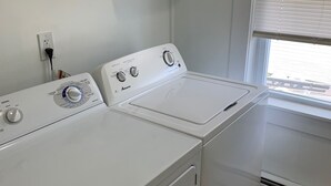 A laundry room with two white front-loading washers and dryers, a white cabinet above the dryers, and a window with blinds. - Ocean view 2 bedroom apartment (Old Orchard Beach)