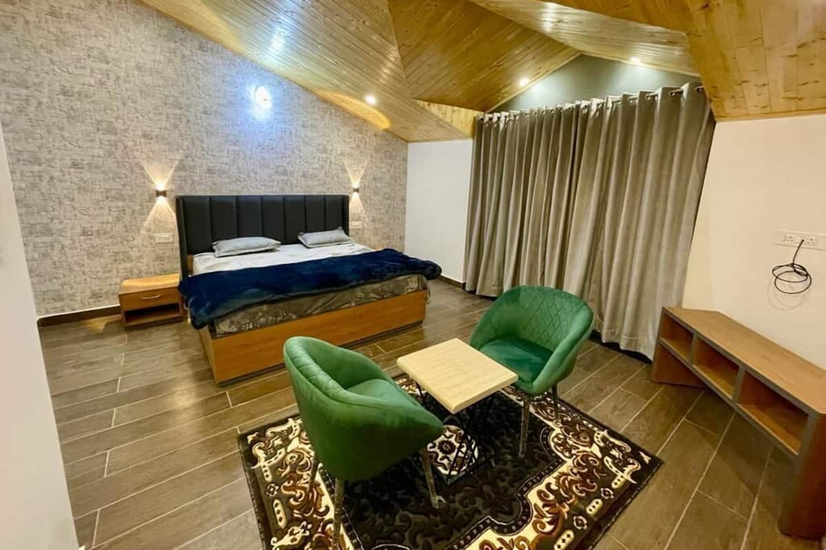 Un dormitorio con una cama, dos sillas, una mesita pequeña, un banco de madera, una alfombra estampada y un techo con vigas de madera.