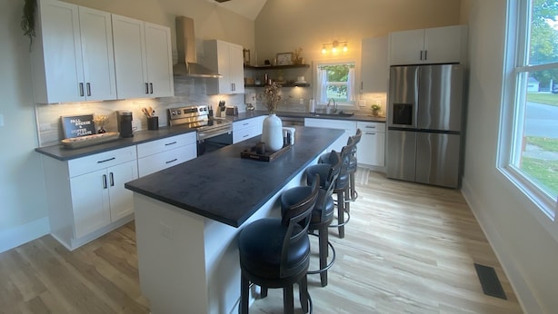 Cozinha moderna com armários brancos, ilha central com banquetas, eletrodomésticos de aço inoxidável e uma grande janela que oferece luz natural.
