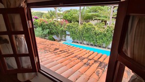 On aperçoit, à travers une fenêtre ouverte, un toit recouvert de tuiles, une piscine et une végétation luxuriante.