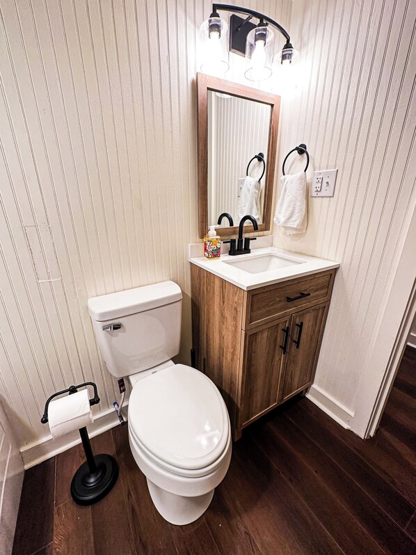 A bathroom with a toilet, sink, wooden cabinet, and a mirror with lights above.