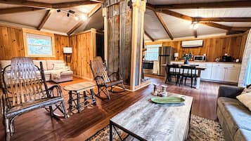 Interior acogedor de una cabaña con paredes de madera, una sala de estar con sofá, mecedoras, una mesa de centro y una zona de cocina con refrigerador y microondas.
