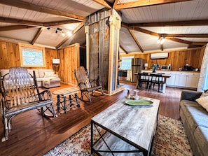 A cozy cabin interior with wooden walls, a living room with a sofa, rocking chairs, a coffee table, and a kitchen area with a refrigerator and microwave. - Blue Door Bungalow of Highlands (Highlands)