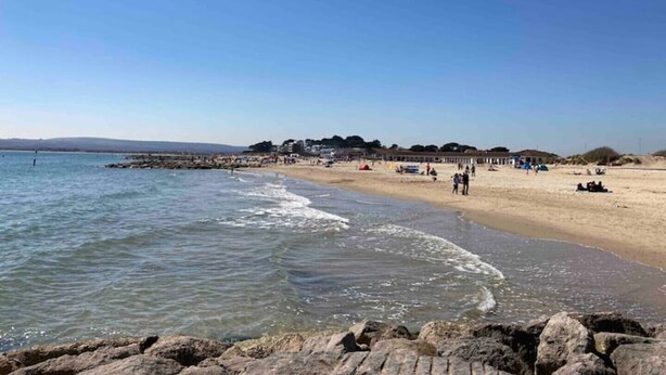 Una playa con cielos despejados, costa rocosa y edificios al fondo.