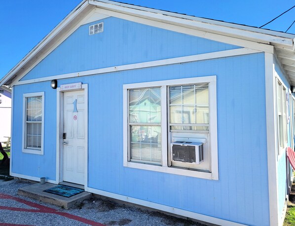 A small, single-story building with a blue exterior, a white door, and two windows with air conditioning units.