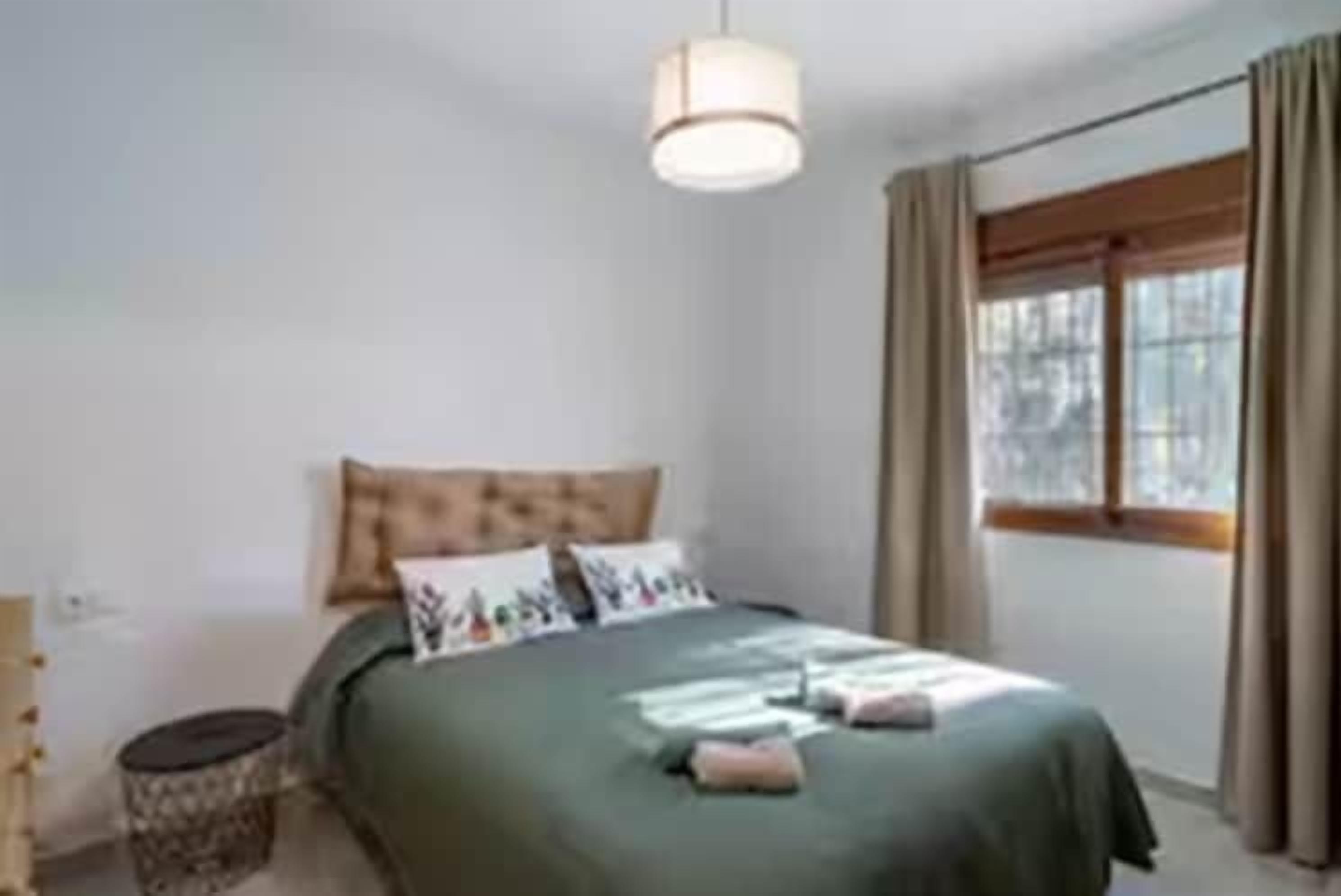 Ein Schlafzimmer mit Bett, Nachttisch, Lampe und einem Fenster mit Vorhängen.