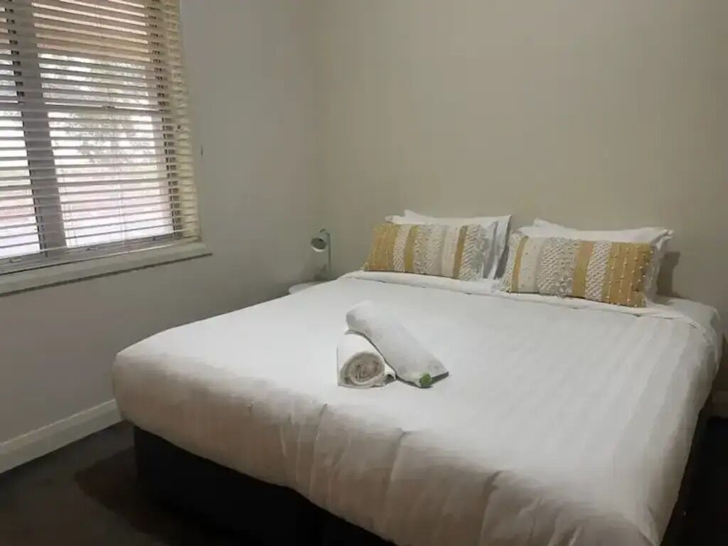 A neatly made bed with white bedding and a rolled towel, a bedside lamp, and a window with blinds.