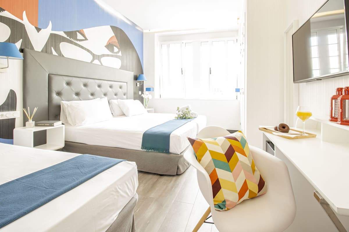 A hotel room with a large bed, a desk, and a chair. There is a TV on the wall and a window with curtains.