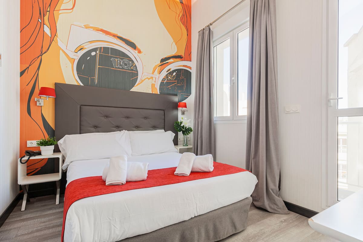 A hotel room with a large bed, a nightstand, a red throw blanket, and a wall mural.