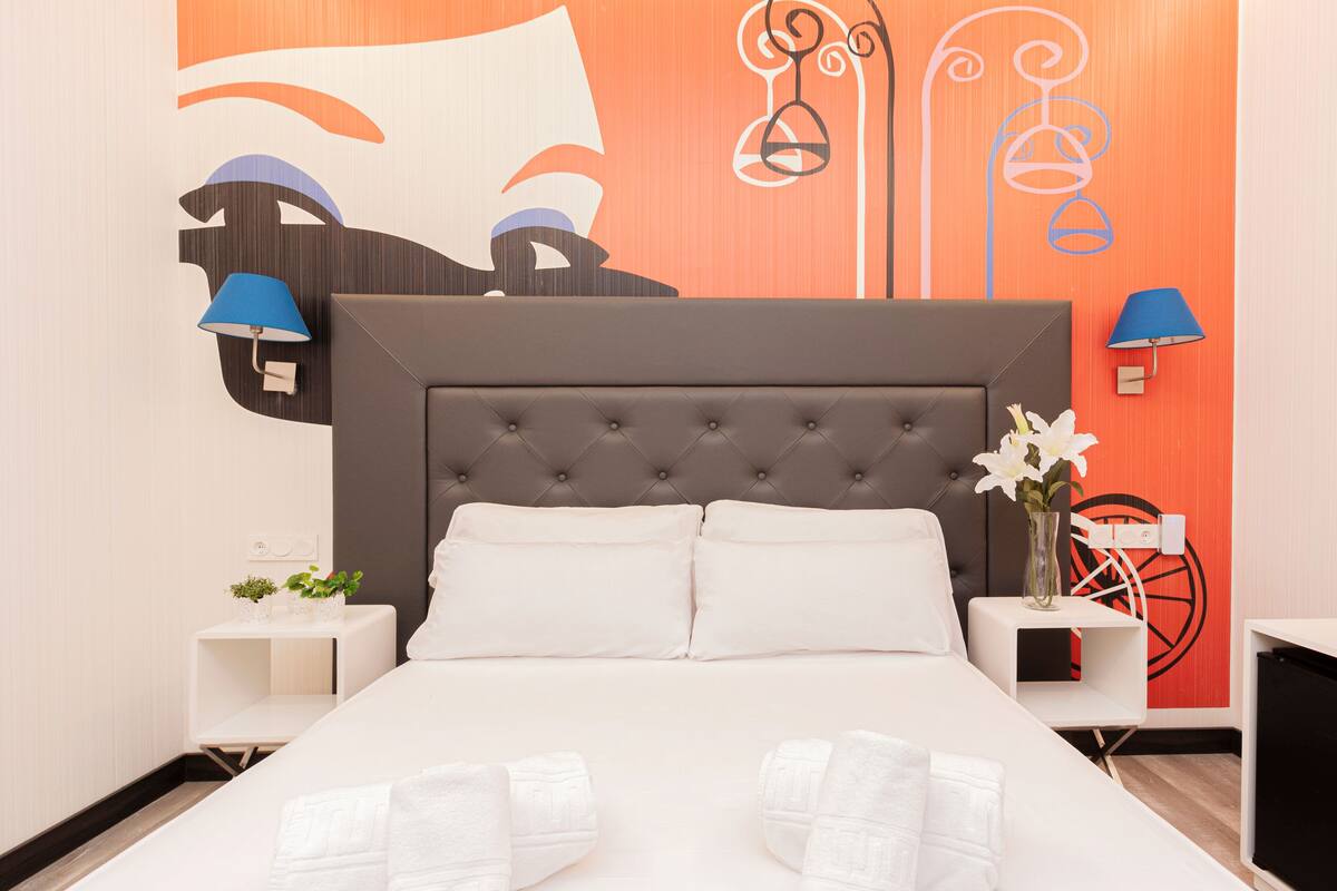 A neatly made bed with white linens and two pillows, a tufted headboard, and a nightstand with a lamp and a vase with flowers.