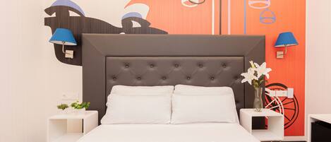 Una cama bien tendida con sábanas blancas y dos almohadas, un cabecero capitonado y una mesita de noche con una lámpara y un florero con flores.