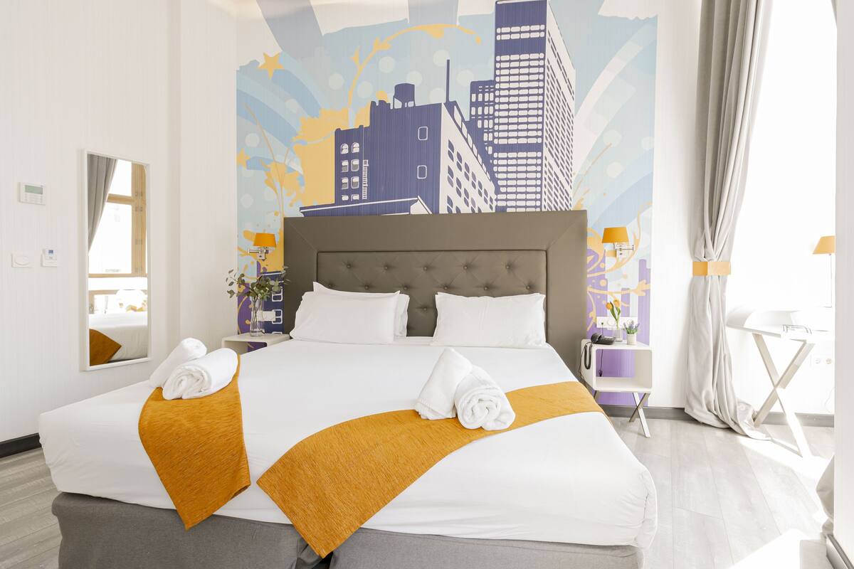 A hotel room with a large bed, a cityscape mural on the wall, a side table with a lamp, and a window with curtains.