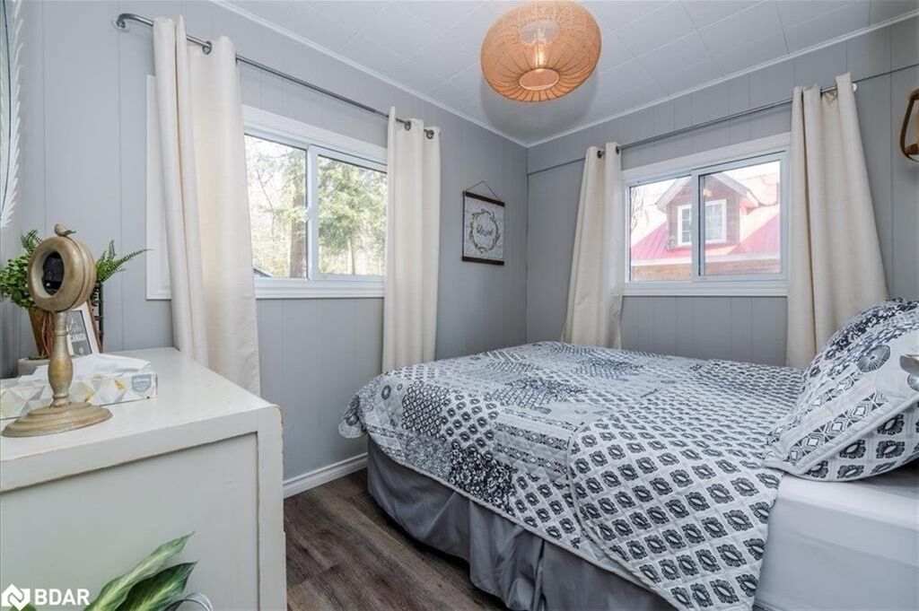 A bedroom with a bed, two windows with curtains, a bedside table with a lamp, and a wall-mounted picture.