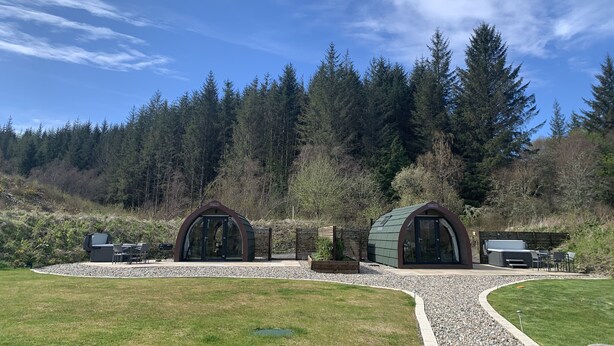 Two modern, semi-circular structures with glass doors, surrounded by a gravel path and a grassy area, set against a backdrop of dense forest and a clear blue sky. - Tawny Owl Pod with Hot Tub (Glencruitten)