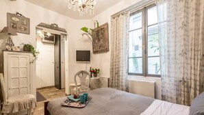 Un dormitorio con cama, una silla, un escritorio, un televisor y una ventana con cortinas.