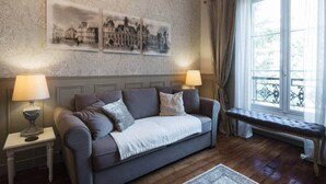Ein gemütliches Wohnzimmer mit einem grauen Sofa, zwei Beistelltischen mit Lampen, einer Bank und einem großen Fenster mit Vorhängen.