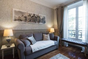 Una sala de estar acogedora con un sofá gris, dos mesas auxiliares con lámparas, un banco y un ventanal con cortinas.