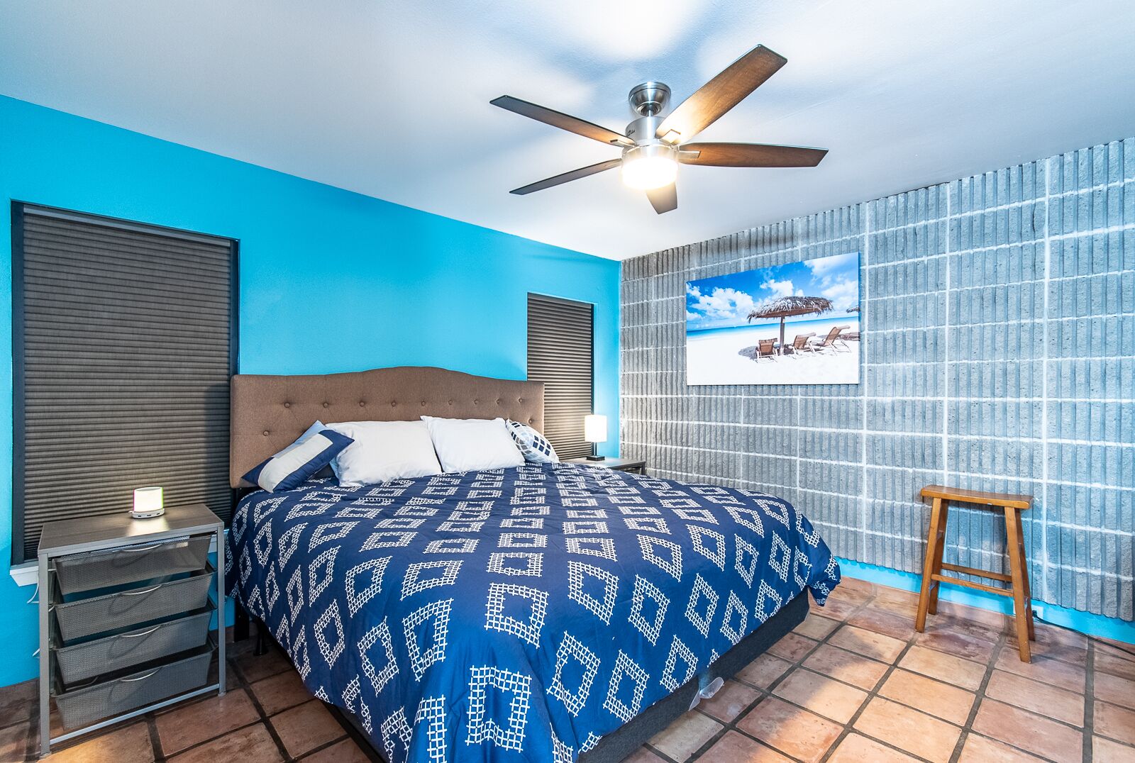 A bedroom with a bed, a nightstand, a ceiling fan, a wall-mounted TV, and a beach-themed painting.