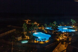 Vue nocturne d’un complexe hôtelier doté d’une grande piscine, entourée de palmiers et bordée de passerelles en bois.