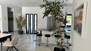 Un comedor moderno con una mesa grande, sillas y taburetes de bar. Hay plantas en macetas y un ventanal con persianas.