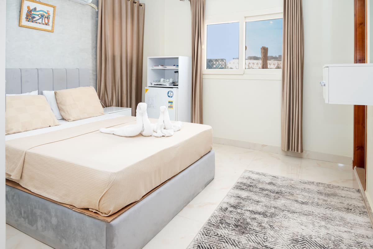 A neatly made bed with a beige comforter and pillows, a small white refrigerator, a window with a view of a fence and trees, and a patterned rug on the floor.