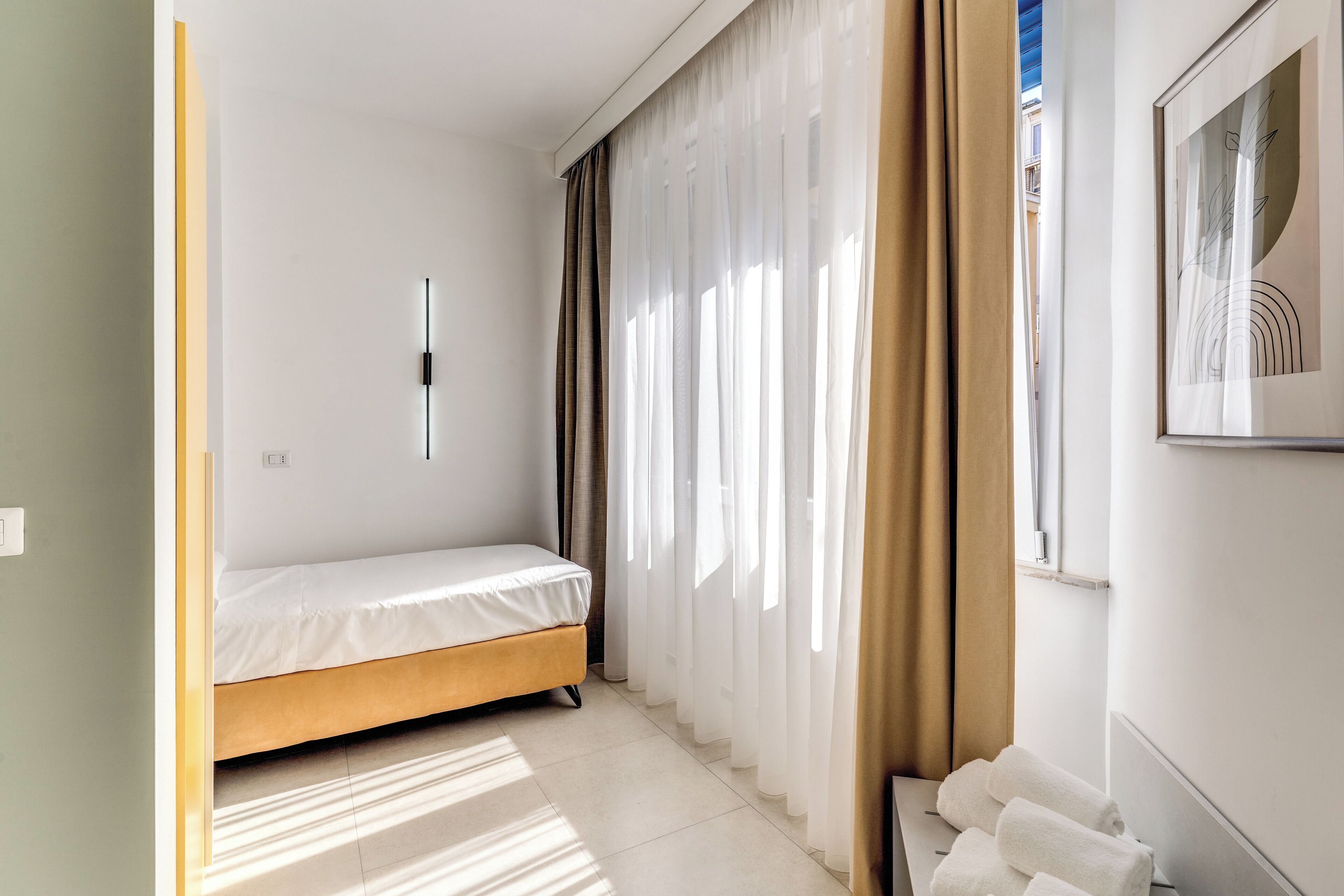 Une petite pièce bien éclairée, avec un lit simple, une fenêtre ornée de rideaux légers et une œuvre d’art encadrée accrochée au mur.