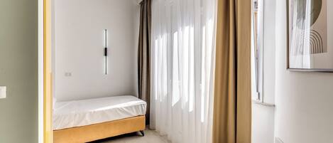 Une petite pièce bien éclairée, avec un lit simple, une fenêtre ornée de rideaux légers et une œuvre d’art encadrée accrochée au mur.