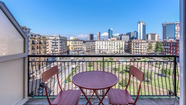 A balcony with two red chairs and a round table, overlooking a cityscape with various buildings and a clear sky. - National Square Napoli (Naples)