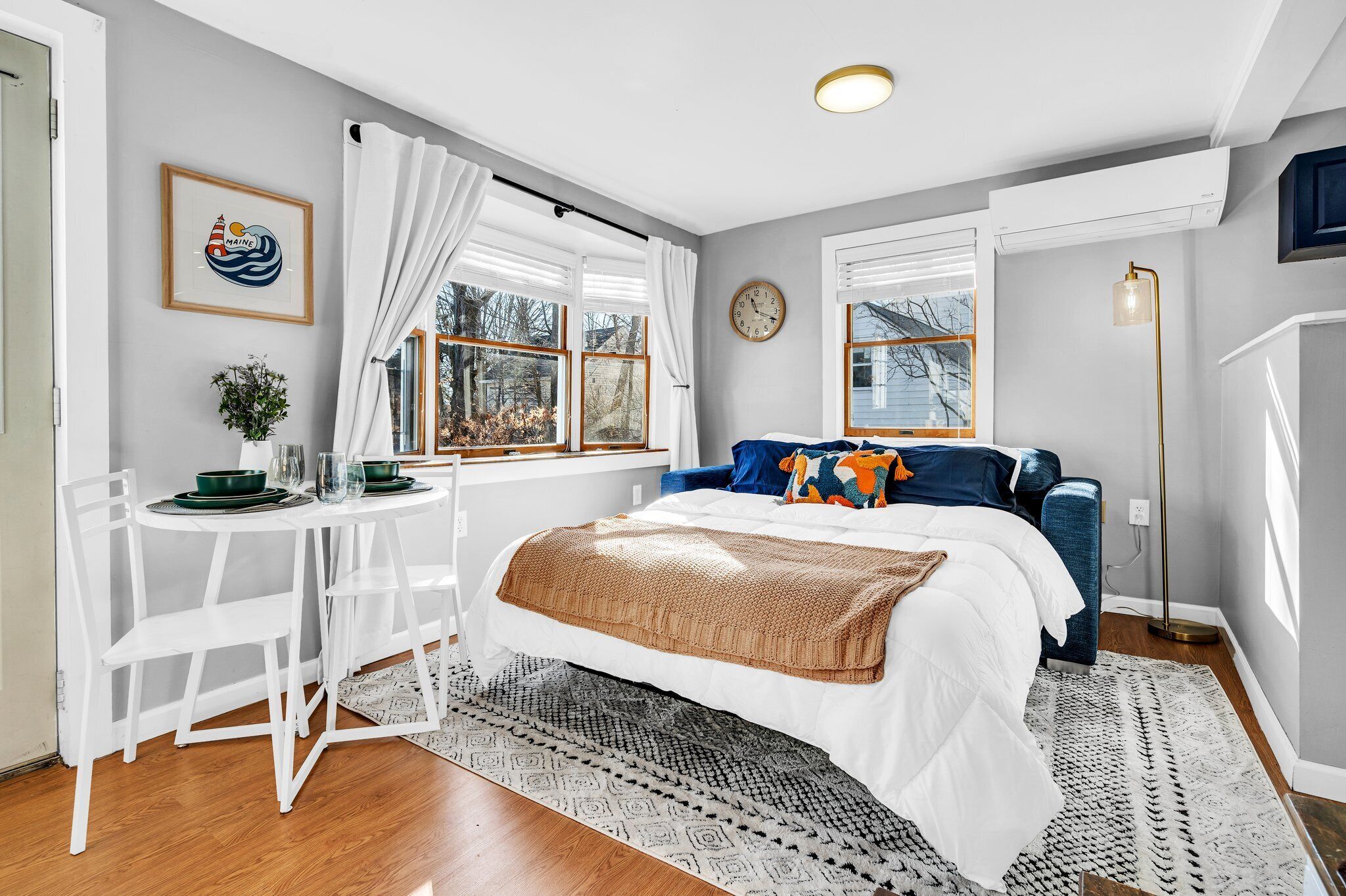 A bedroom with a bed, a desk, and a chair. There is a window with curtains, a clock on the wall, and a plant on the desk.