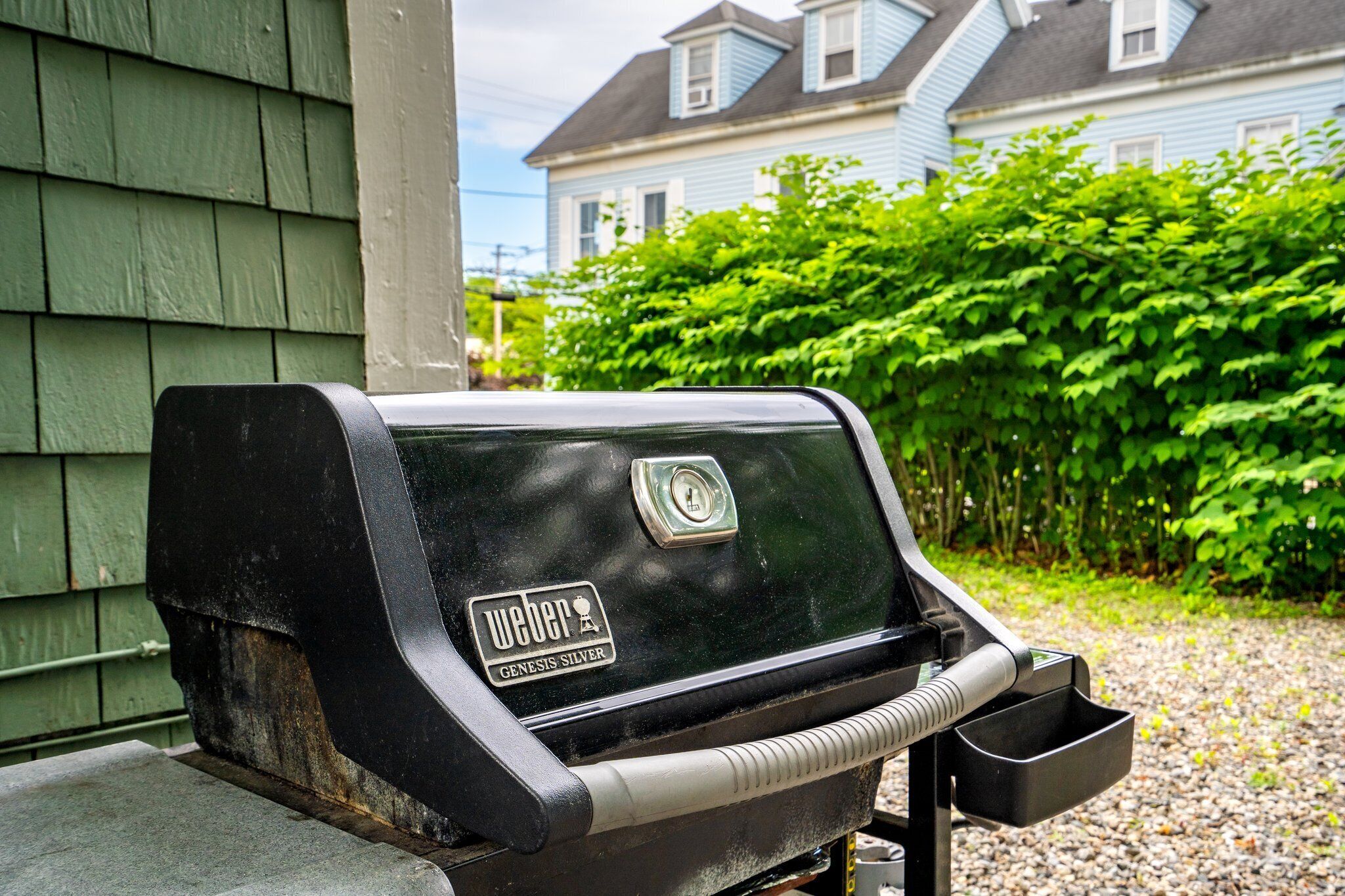 A Weber Genesis Silver grill with a side table, positioned outdoors with a house and greenery in the background.