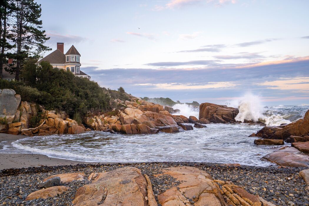 A coastal house with a prominent turret, situated on a rocky shoreline with waves crashing against the rocks.